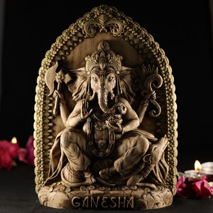 May include: A wooden sculpture of the Hindu deity Ganesha, carved in a detailed style. The sculpture depicts Ganesha sitting on a throne with his elephant head and multiple arms. The sculpture is painted in shades of brown and gold, and the base is inscribed with the word "GANESHA".
