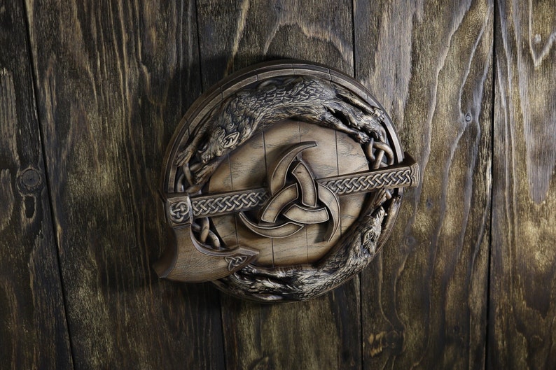 May include: A wooden wall hanging featuring a circular shield with a carved wolf design, a Viking axe, and a triquetra symbol. The shield is brown and the axe is a dark brown color.