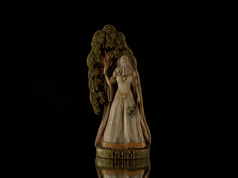 Puede incluir: Una figurita de madera de una mujer con un vestido blanco y velo, sosteniendo una gavilla de trigo. Ella est&aacute; de pie frente a un &aacute;rbol tallado con las palabras "FRANDS FISKUD" en la base.