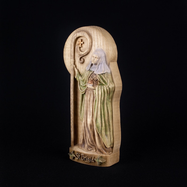 May include: A wooden carving of Saint Brigid, a female saint, holding a staff and a heart. The carving is on a wooden plaque with the words "St Brigid" inscribed below.