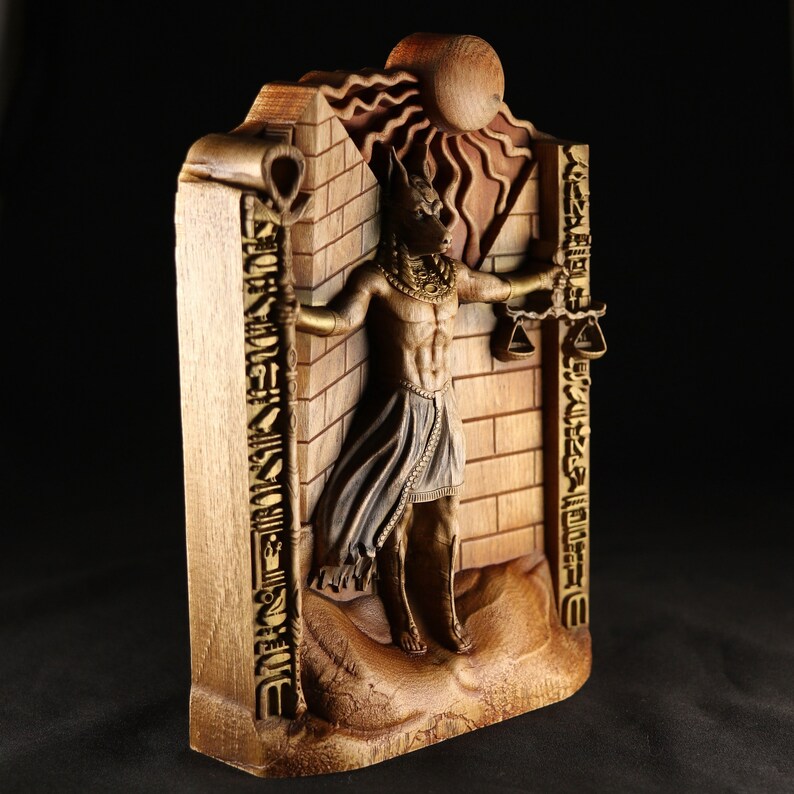May include: A wooden wall sculpture depicting an Egyptian god with a jackal head, holding scales and a staff. The sculpture is carved with intricate details and painted in gold and brown tones. The background features a sun and hieroglyphs.
