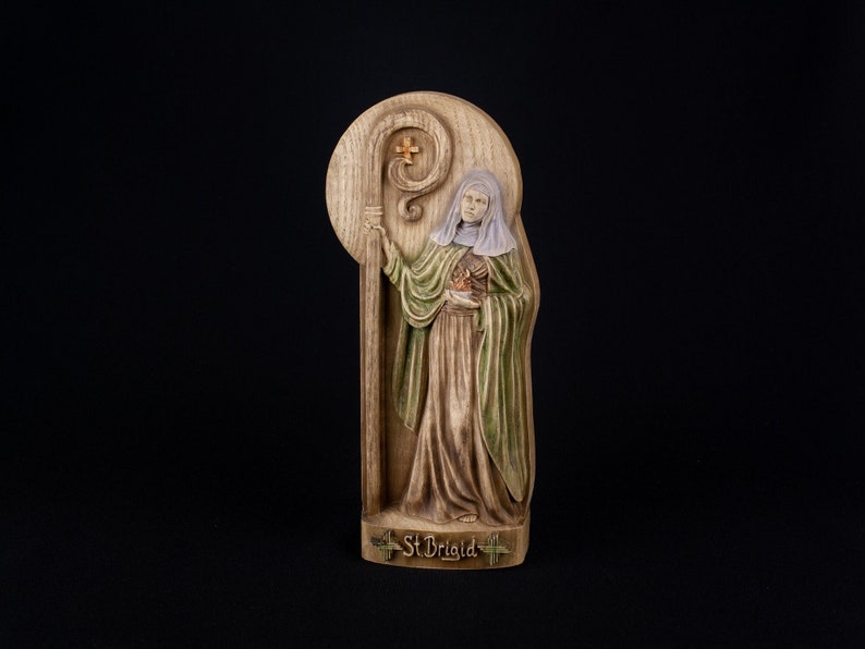 May include: A wooden plaque with a carved image of Saint Brigid, a female saint, holding a staff with a cross. The plaque is painted in shades of brown, green, and white. The text "St Brigid" is written at the bottom of the plaque.