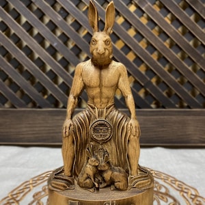 May include: A wooden sculpture of a rabbit-headed humanoid figure sitting with two smaller rabbits at its feet. The figure is carved in a detailed, realistic style and has a muscular physique. The sculpture is painted in a warm brown color and has a smooth, polished finish.