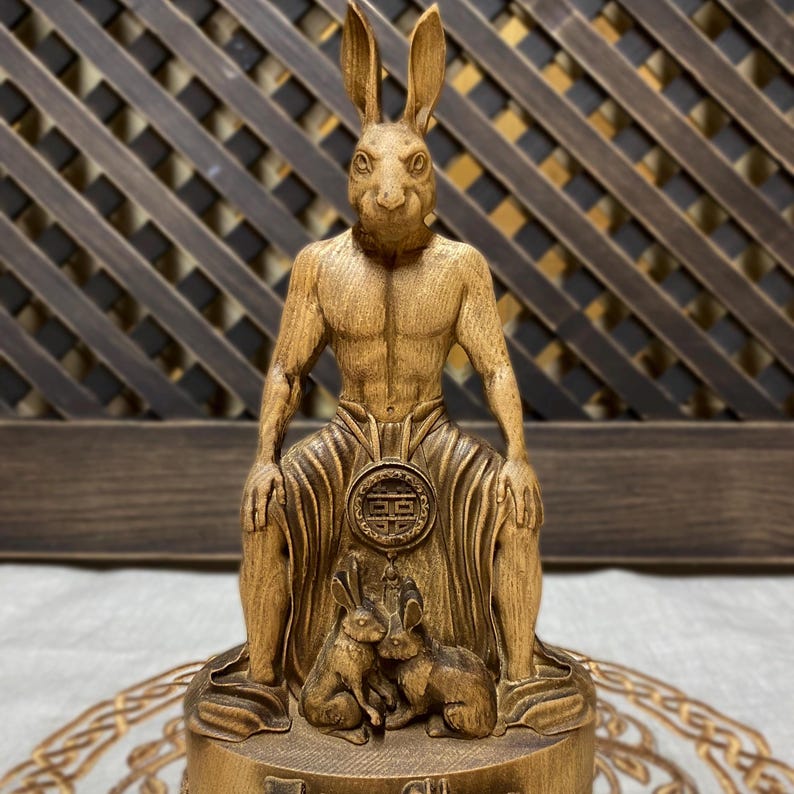 May include: A wooden sculpture of a rabbit-headed humanoid figure sitting with two smaller rabbits at its feet. The figure is carved in a detailed, realistic style and has a muscular physique. The sculpture is painted in a warm brown color and has a smooth, polished finish.