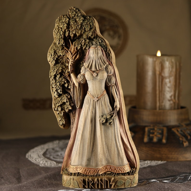 Puede incluir: Una figurita de madera tallada de una mujer con un vestido blanco y velo, de pie frente a un &aacute;rbol. La figurita est&aacute; pintada en tonos marrones y dorados. La palabra "FRIGG" est&aacute; inscrita en la base de la figurita.
