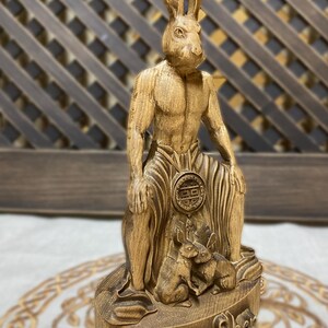 May include: A wooden sculpture of a rabbit-headed humanoid figure sitting with two smaller rabbits at its feet. The sculpture is carved with intricate details and has a rustic, natural finish. The base of the sculpture is inscribed with the words "Twer Shek".