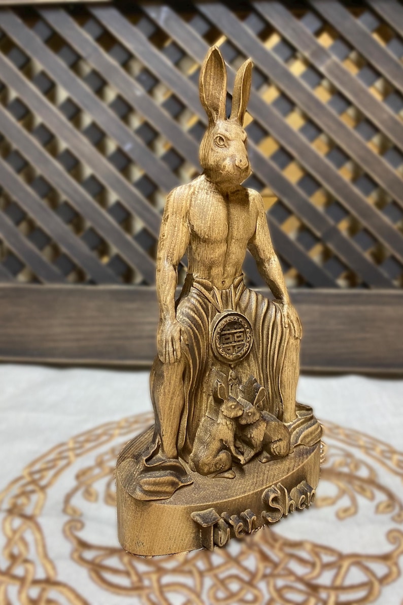 May include: A wooden sculpture of a rabbit-headed humanoid figure sitting with two smaller rabbits at its feet. The sculpture is carved with intricate details and has a rustic, natural finish. The base of the sculpture is inscribed with the words "Twer Shek".