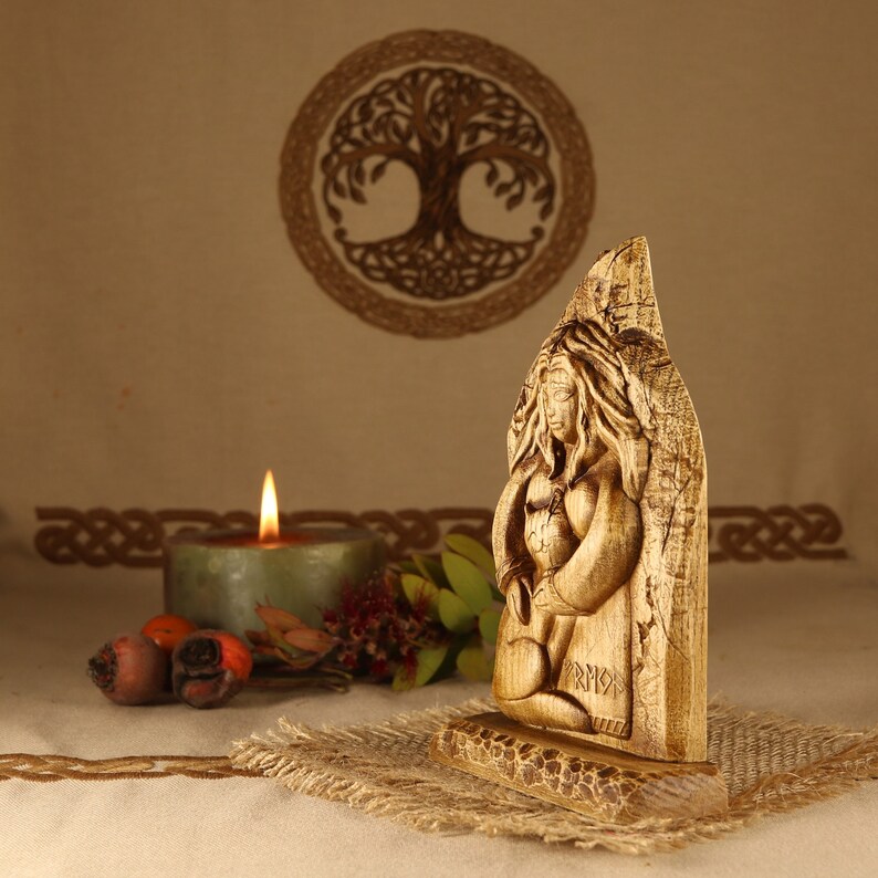 May include: A wooden carving of a Norse goddess, with intricate details and a rustic finish. The carving is set on a wooden base and features the word "FRIGG" carved into the base.