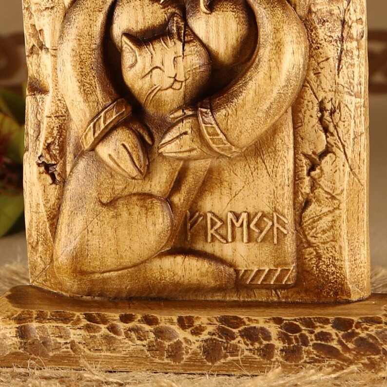 May include: Wooden carving of a cat being held by a person with the word "FRMS" carved below. The carving is on a wooden base.