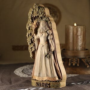 Puede incluir: Una talla de madera de una mujer con un vestido blanco de pie frente a un &aacute;rbol. La talla est&aacute; pintada en tonos marrones y dorados. La mujer sostiene una rama en su mano. La talla est&aacute; etiquetada como "FRIGG" en la parte inferior.