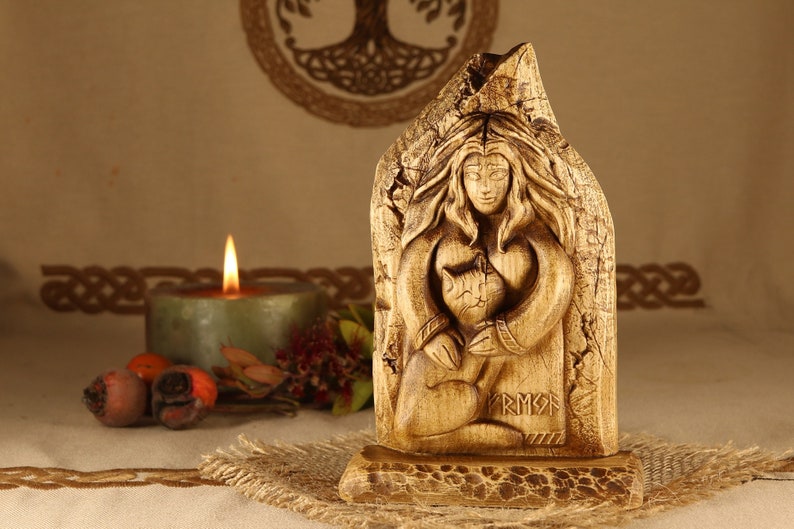 May include: Hand-carved wooden statue of a woman holding a cat, with the inscription "FRIGG" at the bottom. The statue is set against a backdrop of a beige fabric with a woven design.