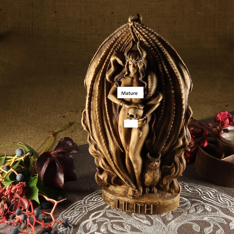 May include: A wooden statue of Lilith, a figure from Jewish mythology, depicted as a nude woman with horns and a skull in her hand. She is standing within a carved, flame-like structure. The base of the statue is inscribed with the word "Lilith".