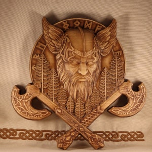 May include: A wooden wall hanging featuring a carved Viking warrior with a horned helmet, surrounded by trees and the word "Odin". Two crossed axes are below the warrior's head.