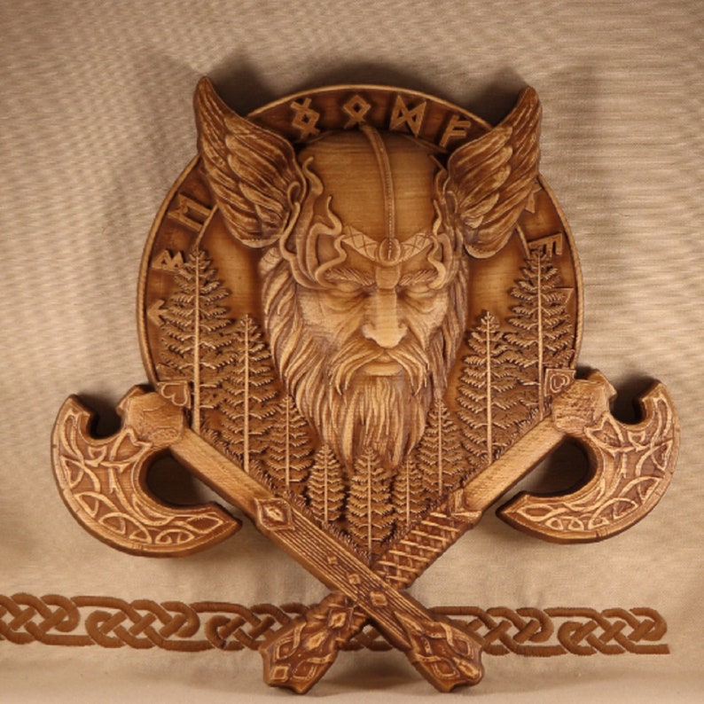 May include: A wooden wall hanging featuring a carved Viking warrior with a horned helmet, surrounded by trees and the word "Odin". Two crossed axes are below the warrior's head.