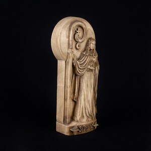 May include: A wooden carving of Saint Brigid, a female saint, holding a staff with a crook. The carving is on a wooden plaque with the words "St. Brigid" inscribed at the bottom.