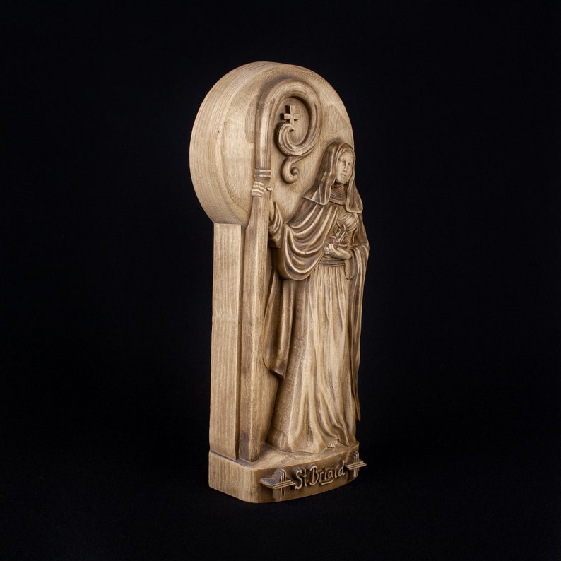 May include: A wooden carving of Saint Brigid, a female saint, holding a staff with a crook. The carving is on a wooden plaque with the words "St. Brigid" inscribed at the bottom.