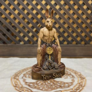 May include: A wooden statue of a rabbit-like deity, Tu'er Shen, sitting with two smaller rabbits at its feet. The statue is painted in shades of brown and gold, with a red and gold robe.
