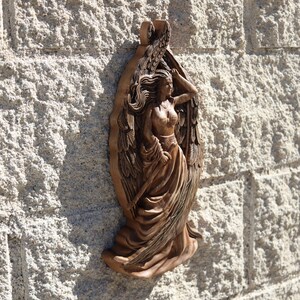 May include: A wooden wall sculpture of a woman with wings. The sculpture is carved in detail and has a natural wood finish.