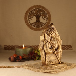 May include: Wooden carving of a bearded figure holding a spear, standing on a base with the inscription "Odin" in runes. The carving is in front of a candle and a woven background.