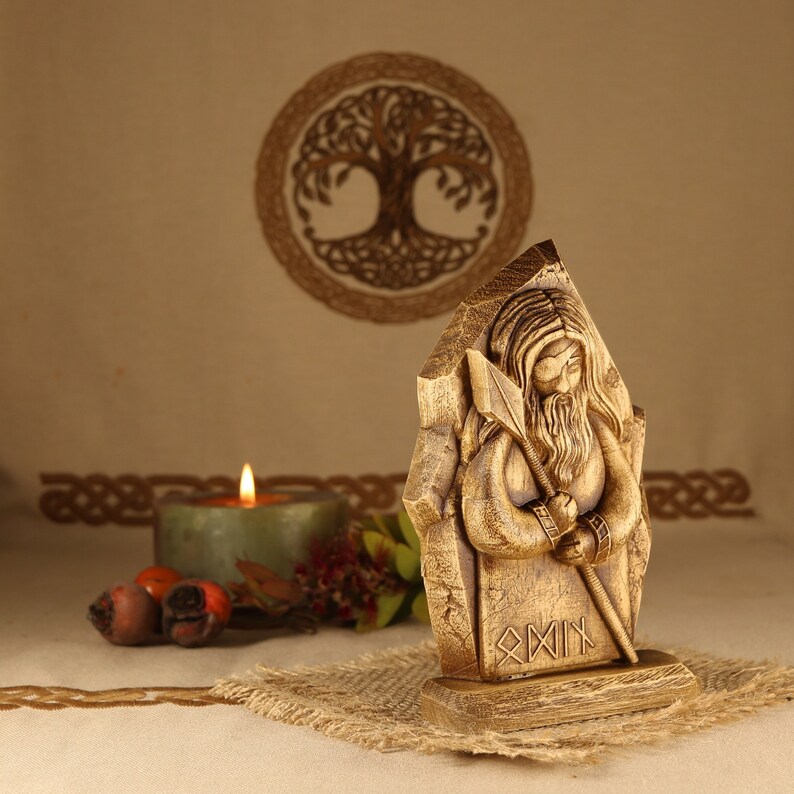 May include: Wooden carving of a bearded figure holding a spear, standing on a base with the inscription "Odin" in runes. The carving is in front of a candle and a woven background.