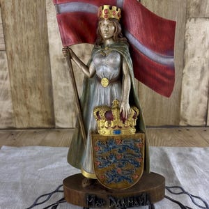 May include: A detailed wooden statue of a woman holding a Danish flag. She wears a crown, a flowing gown, and a cape. A shield with a crown and three lions is at her feet. The base reads "Mor Danmark."