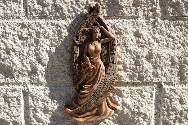 May include: A wooden sculpture of a winged woman with flowing hair and a sword. The sculpture is painted in shades of brown and is mounted on a wall.