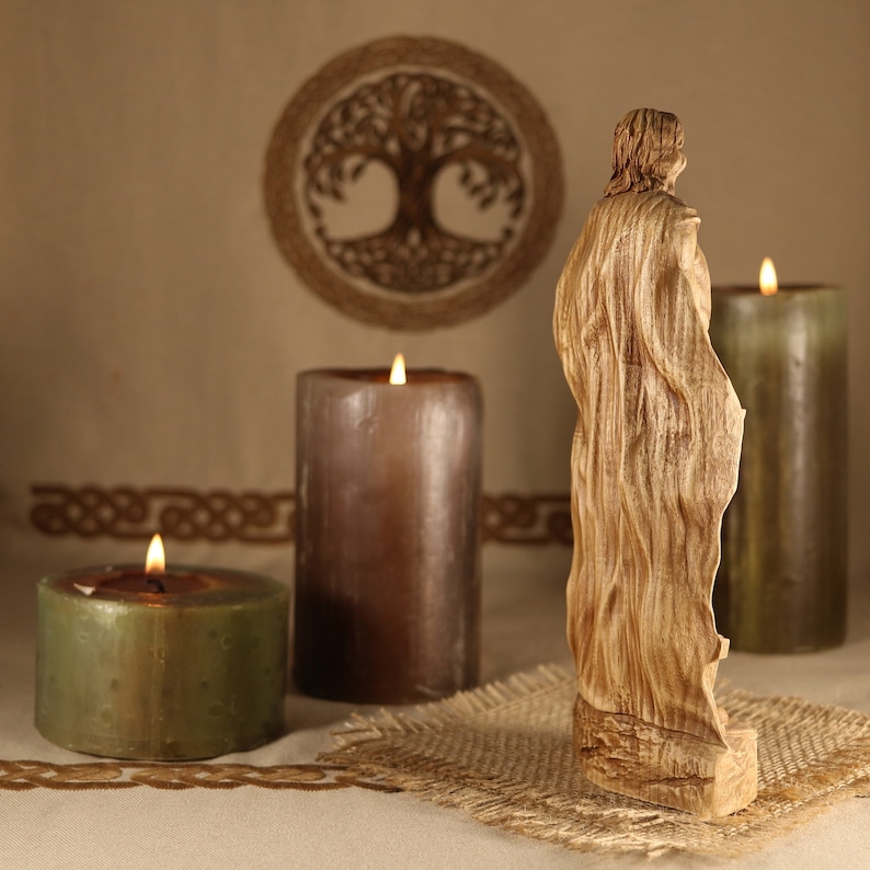 May include: A wooden sculpture of a person standing in front of three candles. The candles are lit and the sculpture is carved with intricate details. The background is a beige fabric with a woven design.