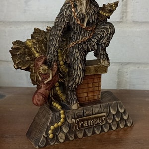 May include: A Krampus figurine, a horned figure with a fur coat and a chain, holding a sack and a child. The figurine is on a base with the name "Krampus" and a chimney. The colors are brown, tan, and gold.