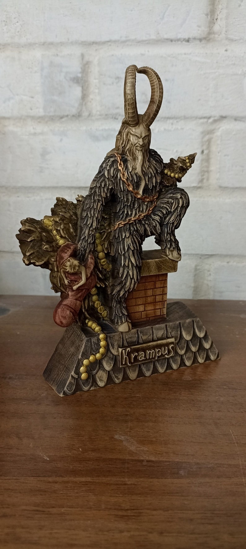 May include: A Krampus figurine, a horned figure with a fur coat and a chain, holding a sack and a child. The figurine is on a base with the name "Krampus" and a chimney. The colors are brown, tan, and gold.