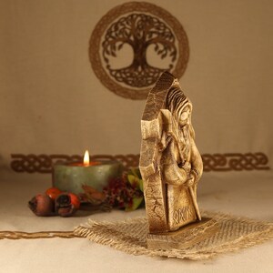 May include: A wooden carving of a bearded figure with a staff, standing in front of a tree of life symbol. The figure is wearing a long robe and has a hood. The carving is on a wooden base with runes carved into it.