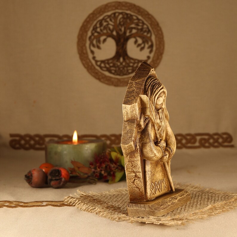 May include: A wooden carving of a bearded figure with a staff, standing in front of a tree of life symbol. The figure is wearing a long robe and has a hood. The carving is on a wooden base with runes carved into it.
