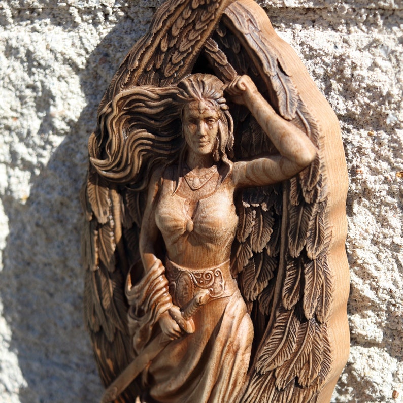May include: A wooden carving of a woman with wings, holding a sword. The woman is wearing a flowing dress and has long hair. The carving is detailed and intricate.