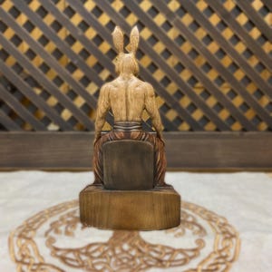 May include: Wooden figurine of a rabbit-headed person sitting on a throne. The figurine is carved in a realistic style and has a detailed design. The figurine is painted in shades of brown and beige.