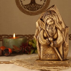 May include: A wooden statue of a bearded figure holding a spear, with the inscription "Odin" in runes below. The statue is carved in a rustic style and has a golden brown finish.