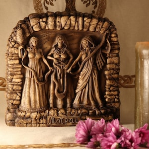 May include: A brown wooden plaque with a relief carving of three figures, possibly goddesses, holding hands. The plaque is set against a beige background with a lit candle and pink flowers in the foreground. The plaque is inscribed with the word "Morrigan".