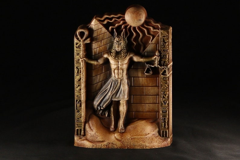 May include: A wooden wall sculpture depicting an Egyptian god with a jackal head. The god is holding a staff with an ankh symbol and a scale in his other hand. The sculpture is decorated with hieroglyphs and a sun disc.