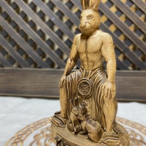 May include: A wooden statue of a rabbit-human hybrid, known as the "Tuer Shen", sitting on a throne with two smaller rabbits at its feet. The statue is carved in a detailed and realistic style, with a warm brown color.