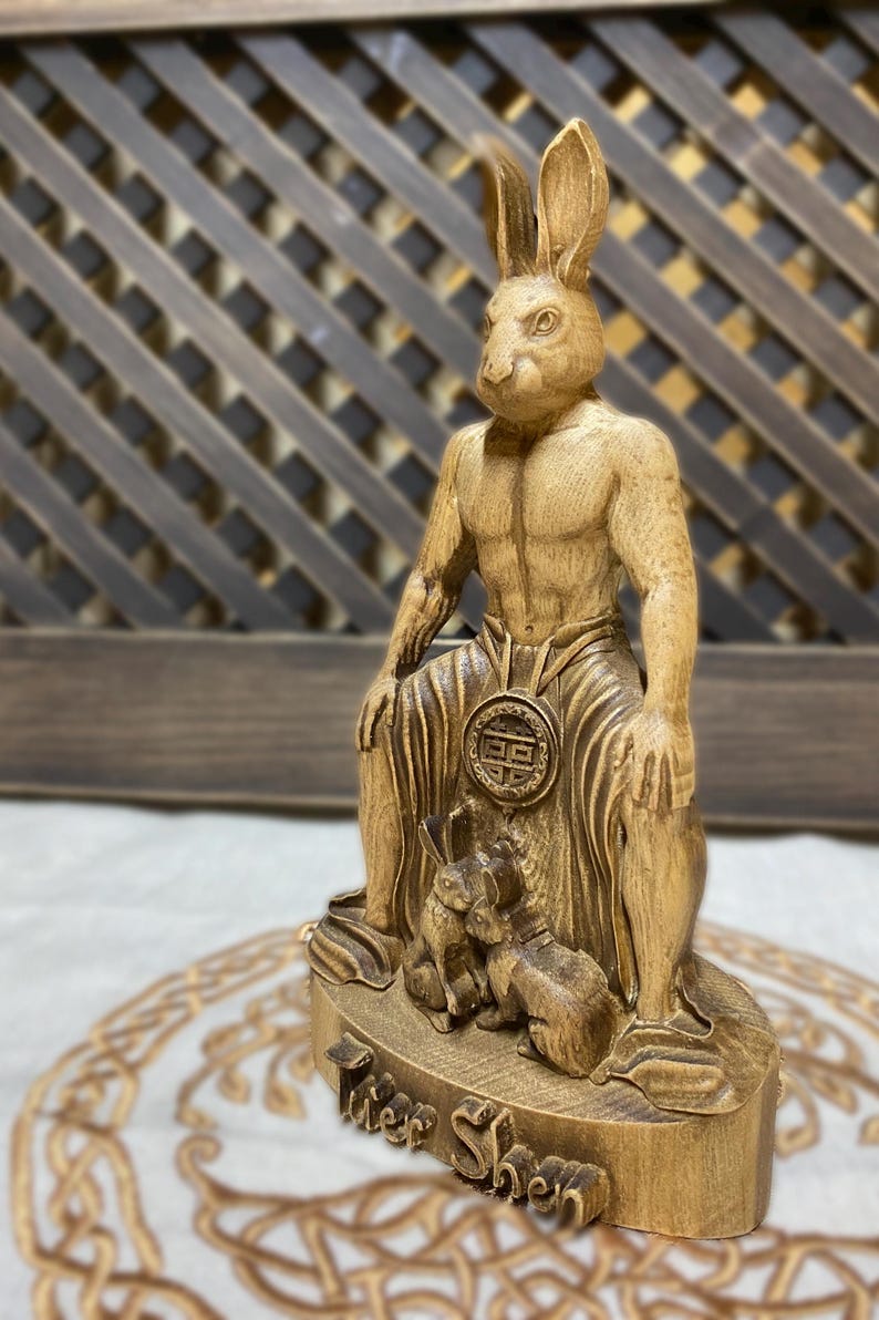 May include: A wooden statue of a rabbit-human hybrid, known as the "Tuer Shen", sitting on a throne with two smaller rabbits at its feet. The statue is carved in a detailed and realistic style, with a warm brown color.