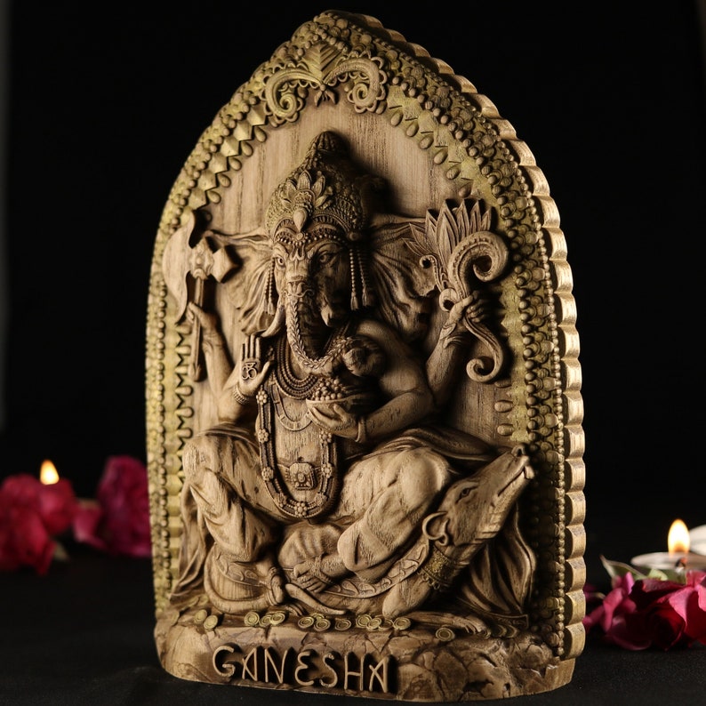 May include: A wooden carving of the Hindu deity Ganesha, with intricate details and a gold-painted frame. The carving depicts Ganesha sitting on a throne with his elephant head and multiple arms.