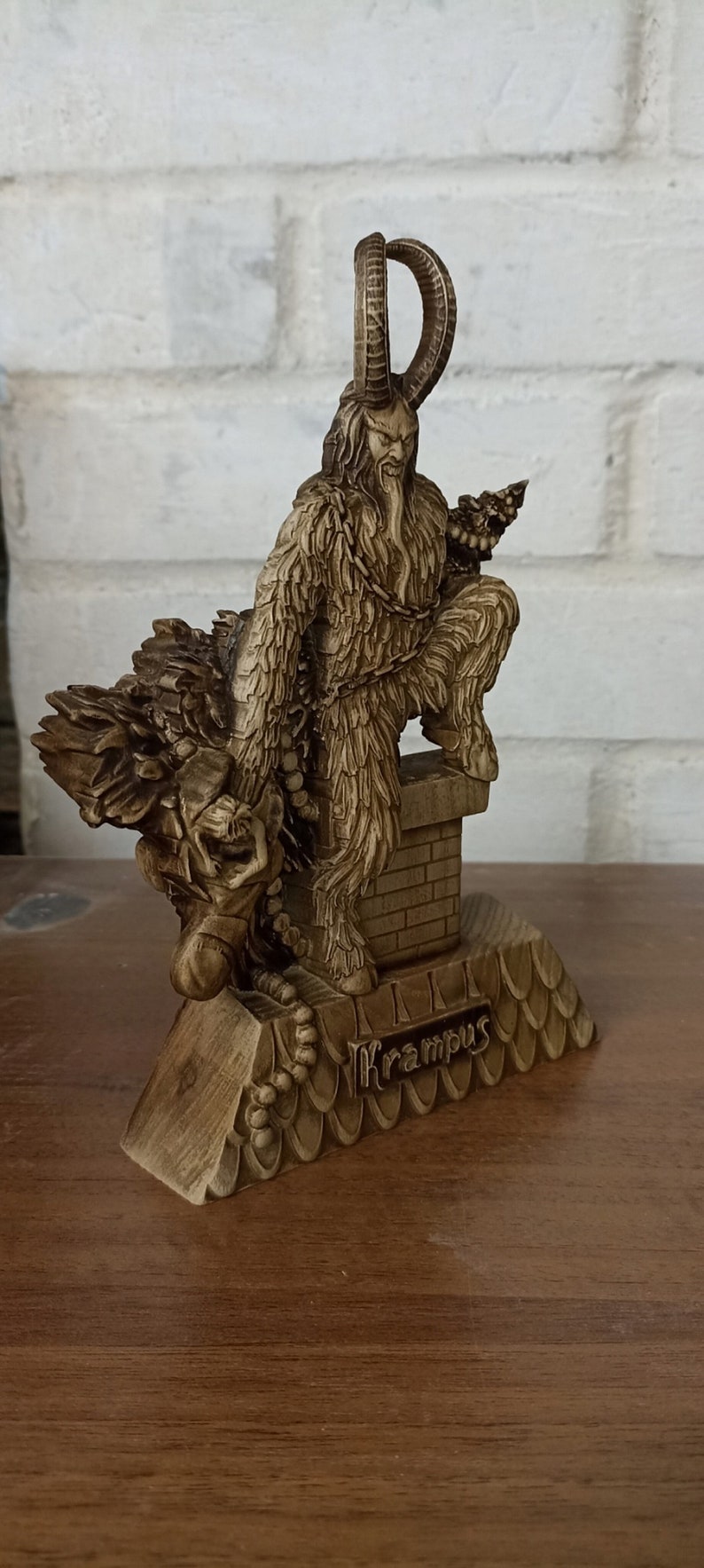 May include: A detailed wooden Krampus figurine, featuring a horned, fur-covered creature with a chain, perched atop a chimney. The figurine is a light brown color and has intricate carvings. The word "Krampus" is inscribed on the base.