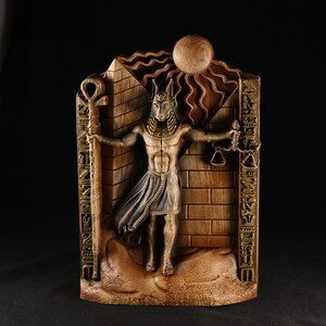 May include: A wooden wall sculpture depicting an Egyptian god with a jackal head, holding a scale and a staff. The sculpture is painted in gold and brown tones and features hieroglyphs.