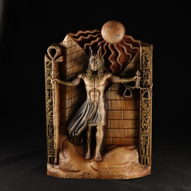May include: A wooden wall sculpture depicting an Egyptian god with a jackal head, holding a scale and a staff. The sculpture is painted in gold and brown tones and features hieroglyphs.