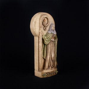 May include: A wooden figurine of Saint Brigid, a female saint, holding a staff with a cross. The figurine is painted in shades of brown, green, and white. The base of the figurine is inscribed with the words "St. Brigid".