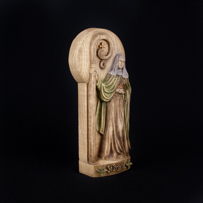 May include: A wooden figurine of Saint Brigid, a female saint, holding a staff with a cross. The figurine is painted in shades of brown, green, and white. The base of the figurine is inscribed with the words "St. Brigid".