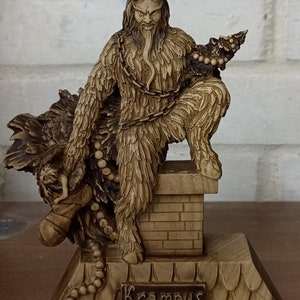 May include: A detailed figurine of Krampus, a horned figure from folklore, with a long tongue and chains. The figurine is brown and sits atop a decorative base with the name "Krampus" on it. The background is a white brick wall.