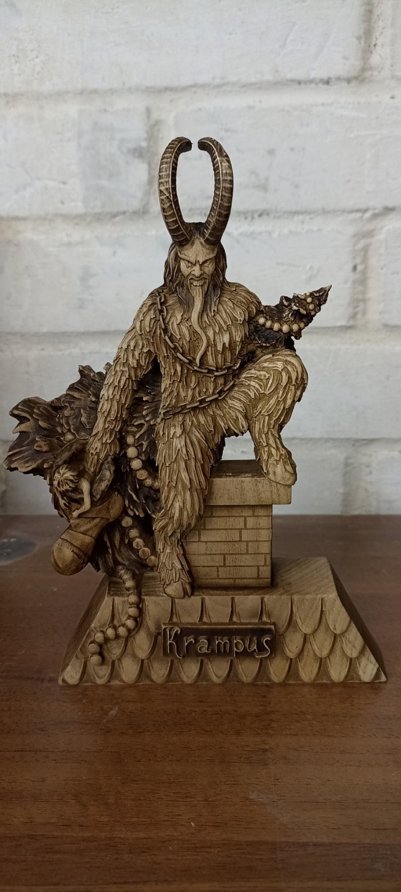 May include: A detailed figurine of Krampus, a horned figure from folklore, with a long tongue and chains. The figurine is brown and sits atop a decorative base with the name "Krampus" on it. The background is a white brick wall.