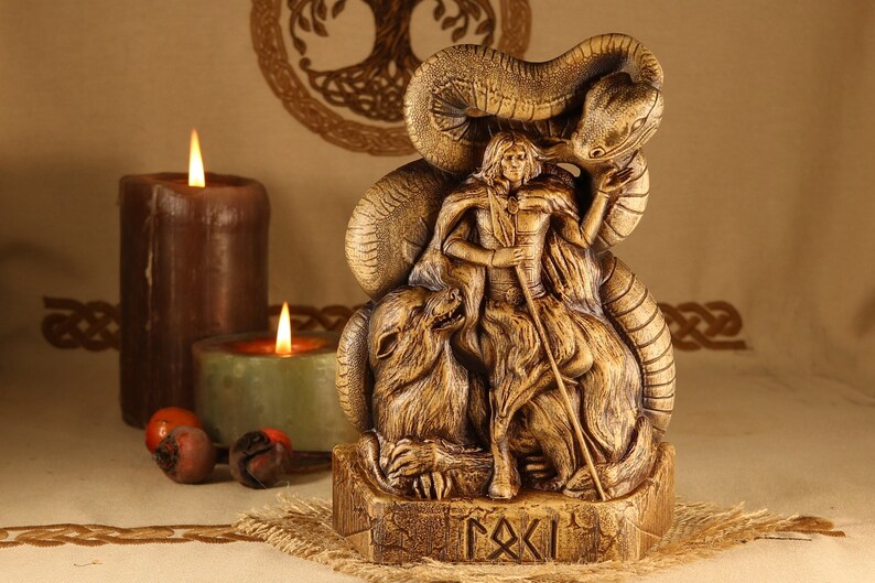 May include: A wooden statue of a Norse god, possibly Odin, with a wolf and a serpent. The statue is brown and gold in color and has intricate details. The base of the statue has the word "RAXI" carved into it.