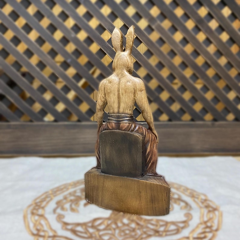 May include: A wooden sculpture of a rabbit-headed figure sitting on a throne. The figure is carved in detail and has a muscular build. The sculpture is painted in shades of brown and beige.