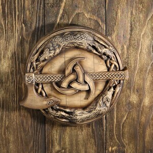 May include: A wooden wall plaque with a carved Viking shield design. The shield features a central triquetra symbol, surrounded by two wolves. An axe is attached to the shield.