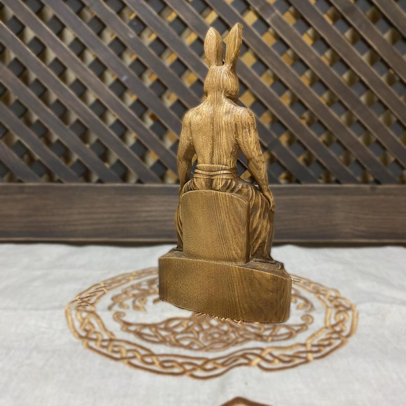 May include: Wooden figurine of a rabbit-like creature sitting on a throne. The figurine is carved from light brown wood and has a detailed design. The figurine is sitting on a wooden base.